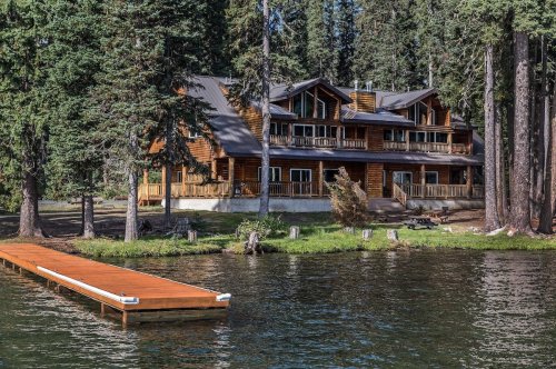 A large wooden cabin by a lake, surrounded by tall trees and featuring a wooden dock in the foreground.