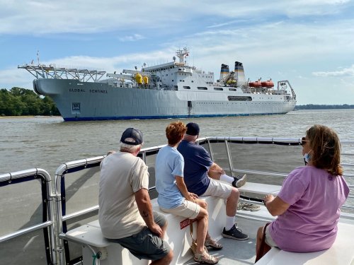A group of four people sits on a boat, watching a large ship, the Global Nominee, pass by on the water.