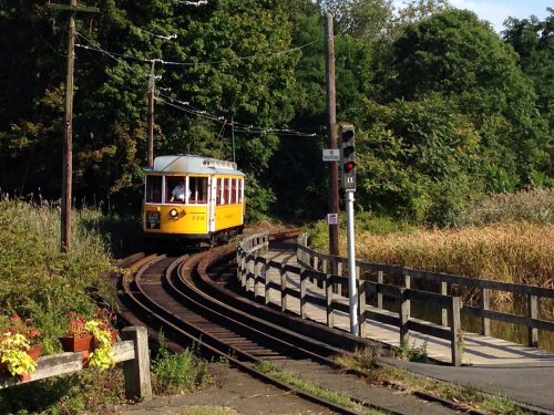 A yellow tram curves along tracks near greenery and a wooden railing, with a signal light visible nearby.