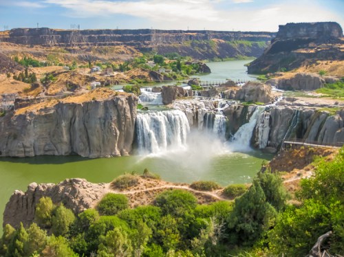 A scenic view of a waterfall cascading into a green river, surrounded by rocky cliffs and lush vegetation.