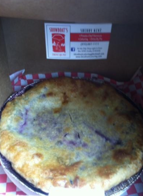 A freshly baked pie in a black tin, resting in a takeout box with a red and white checkered liner.