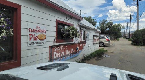 A quaint pie shop with a sign for "Emergency Pie" and colorful flowers, set against a cloudy sky.