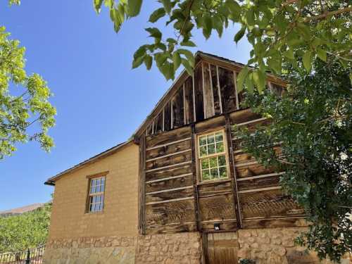 A rustic building with a wooden upper section and stone lower section, surrounded by green trees and a clear blue sky.