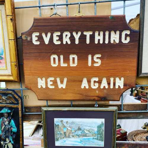 A wooden sign with the phrase "Everything Old Is New Again" displayed at a market or fair.