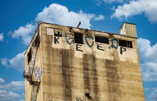 A weathered building with graffiti reading "KEEP" against a backdrop of blue sky and fluffy clouds.