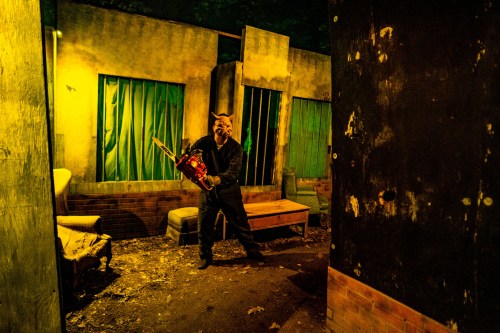 A masked figure holds a chainsaw in a dimly lit, abandoned room with green curtains and scattered debris.