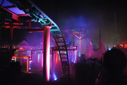 A colorful roller coaster at night, illuminated by vibrant lights and surrounded by mist.