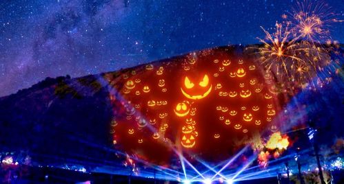 A mountain illuminated with Halloween-themed jack-o'-lanterns, under a starry sky and fireworks.