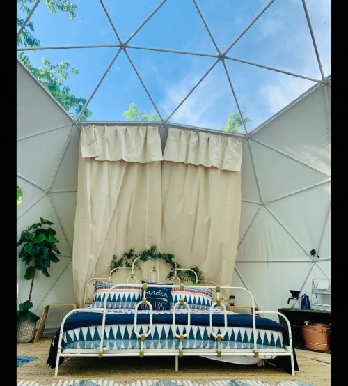 A cozy bedroom inside a geodesic dome, featuring a bed with patterned bedding, a plant, and large windows.