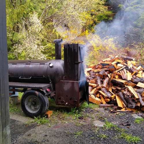 A black smoker next to a pile of firewood, surrounded by lush greenery and misty atmosphere.