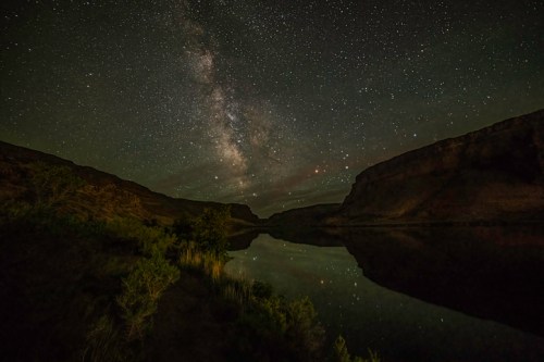 A serene landscape at night featuring a starry sky and the Milky Way reflected in a calm river surrounded by cliffs.