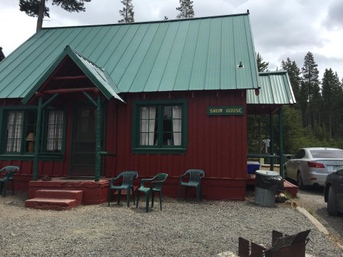 A red cabin with a green roof, labeled "Snow Goose," surrounded by trees and gravel, with chairs and a car nearby.