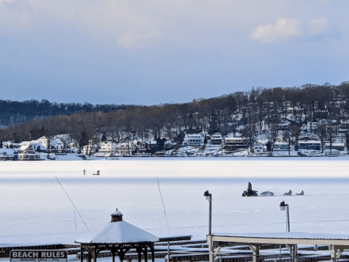 A snowy landscape with a frozen lake, houses in the background, and people ice fishing.