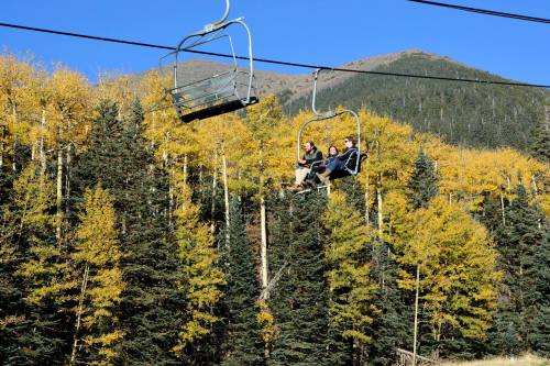 Three people ride a ski lift surrounded by vibrant autumn trees and a mountain backdrop under a clear blue sky.