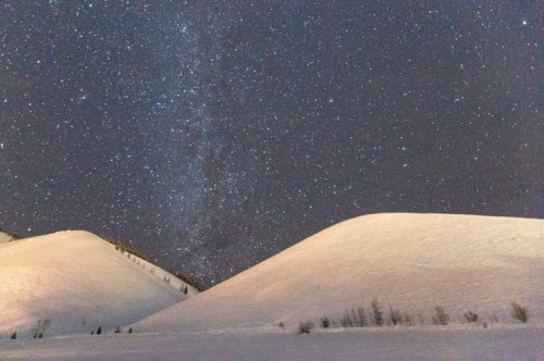 A snowy landscape with two large dunes under a starry night sky, featuring the Milky Way in the background.