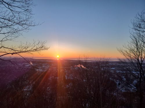 A vibrant sunset over a winter landscape, with trees silhouetted in the foreground and soft clouds in the sky.