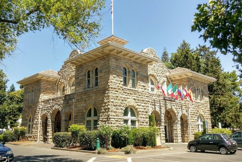 Historic stone building with arched windows, flags from various countries, and surrounded by trees under a clear blue sky.