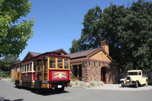 A vintage trolley parked near a stone building and an old-fashioned car under a clear blue sky.