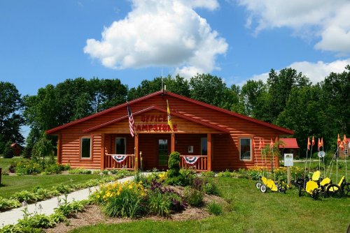 A red log cabin building with flags, surrounded by colorful flowers and greenery under a blue sky with clouds.