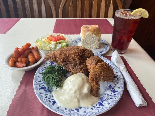 A plate of fried chicken with gravy, carrots, salad, a biscuit, and a glass of iced tea on a table.