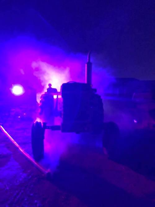 A tractor illuminated by purple and blue lights, surrounded by smoke, creating a dramatic nighttime scene.