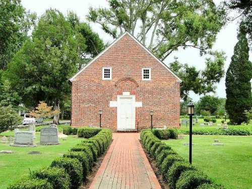 A brick building with a white door, surrounded by neatly trimmed hedges and gravestones in a green cemetery.