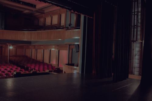 A dimly lit theater stage with red seats visible in the background, curtains partially drawn.