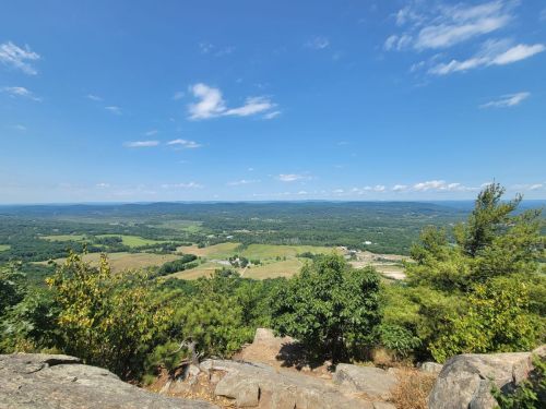 A panoramic view of lush green fields and distant hills under a clear blue sky with scattered clouds.