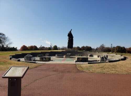 A large statue stands in a circular stone area, surrounded by grass and trees under a clear blue sky.
