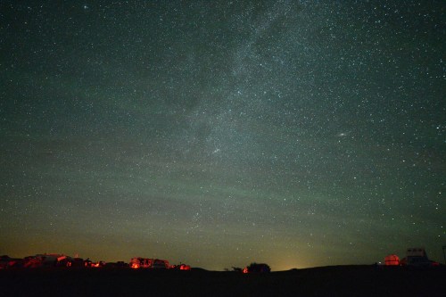 A starry night sky with a faint Milky Way and distant lights from buildings below.