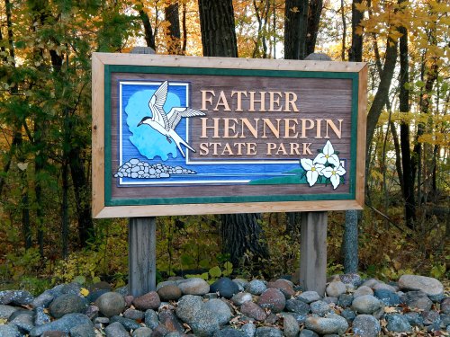 Sign for Father Hennepin State Park, featuring a bird and floral design, surrounded by trees and rocks.