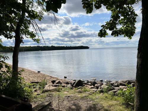 A serene lakeside view with calm water, sandy shore, and trees framing the scene under a partly cloudy sky.