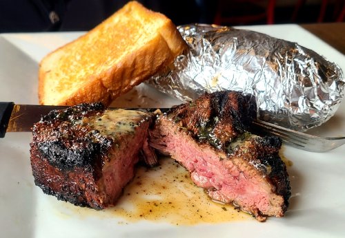 A plate featuring a grilled steak with a buttery glaze, a baked potato wrapped in foil, and a slice of toasted bread.