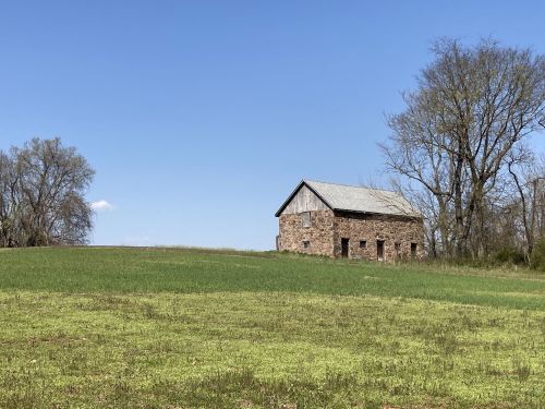 A rustic stone barn sits on a grassy hill under a clear blue sky, surrounded by trees.