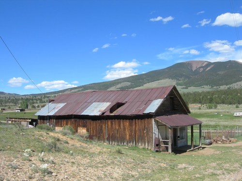 A rustic wooden barn with a metal roof, set against a backdrop of mountains and blue skies.