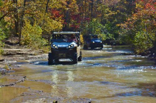 Two off-road vehicles drive through a shallow stream surrounded by colorful autumn foliage.