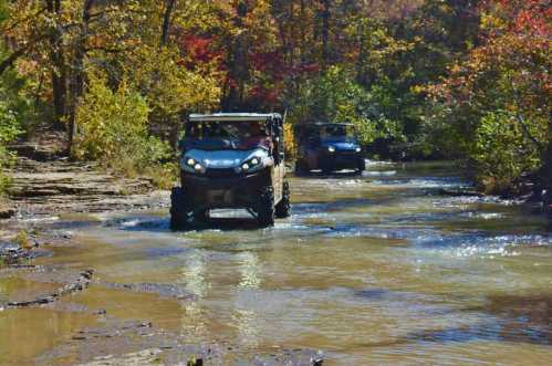 Two off-road vehicles drive through a shallow stream surrounded by colorful autumn foliage.