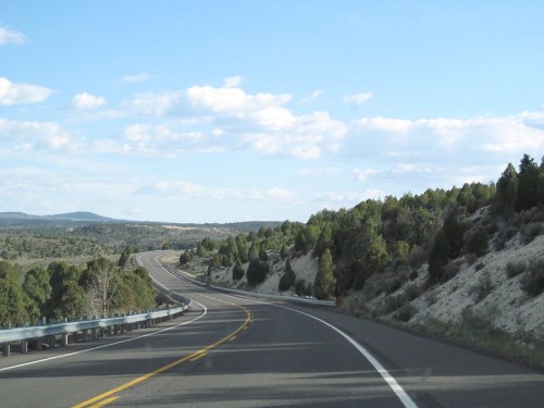 A winding road stretches through a landscape of trees and hills under a blue sky with scattered clouds.