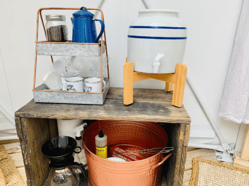 A wooden shelf with a blue kettle, white water dispenser, and various kitchen items, including cups and utensils.