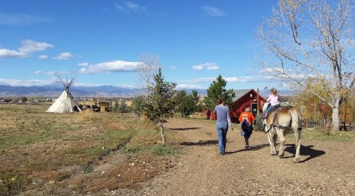A person walks alongside a child on a horse, with a teepee and mountains in the background under a blue sky.