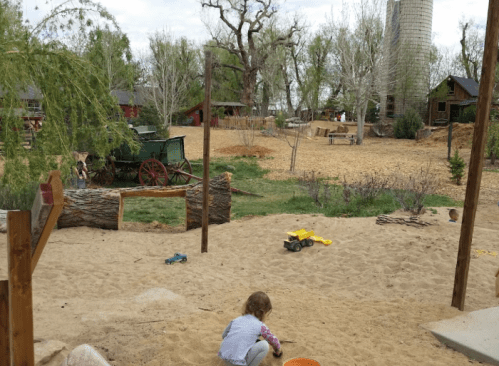 A child plays in a sandy area on a farm, with trees, a barn, and a silo in the background.