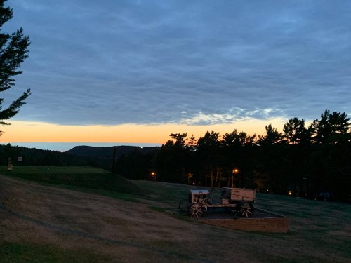 A serene landscape at dusk, featuring a wagon on grassy terrain with trees and a colorful sunset in the background.