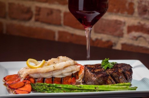 A gourmet plate featuring grilled lobster tail, steak, asparagus, and a glass of red wine in the background.