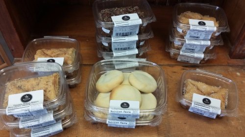 Assorted baked goods in clear containers on a wooden shelf, labeled with nutrition facts.