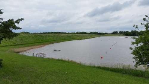 A calm lake surrounded by green grass and trees, with a small dock and buoy markers in the water under a cloudy sky.