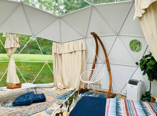 Cozy interior of a geodesic dome with a hanging chair, rugs, and natural light from large windows.