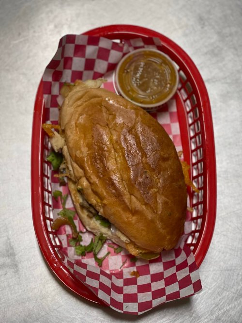 A sandwich in a red basket lined with checkered paper, accompanied by a small cup of sauce.