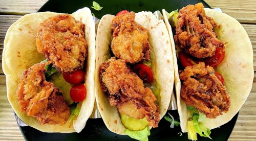Three tacos filled with crispy fried chicken, lettuce, pickles, and cherry tomatoes on a black plate.