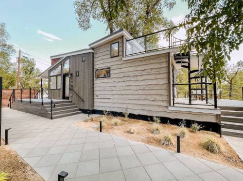 Modern wooden house with large windows, a deck, and a landscaped pathway surrounded by trees.