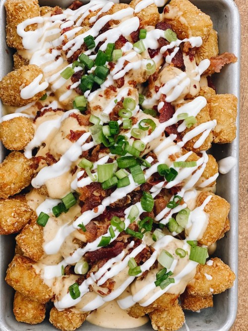A tray of loaded tater tots topped with bacon, green onions, and drizzled with creamy sauce.